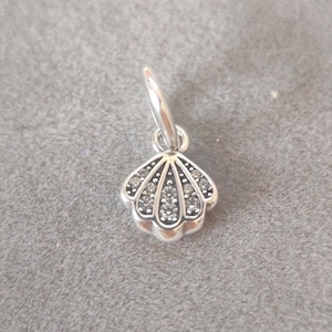 Puede incluir: Un charm de concha plateada con un lazo para pulsera o collar. El charm está detallado con pequeños cristales transparentes y un borde festoneado, creando un efecto brillante. El charm está diseñado para ser un colgante.