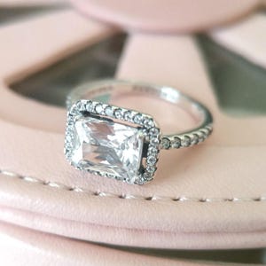 Peut inclure: Une bague en argent avec une grande pierre rectangulaire transparente entourée de petites pierres. La bande de la bague est également décorée de petites pierres.