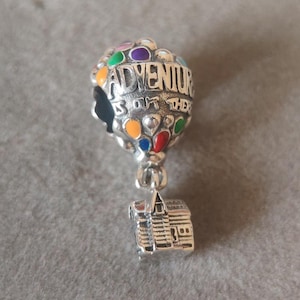 Può includere: Un charm in argento con un design a mongolfiera con accenti colorati e la scritta "ADVENTURE IS OUT THERE". Sotto, un charm dettagliato a forma di casa. Il charm è probabilmente per un braccialetto o una collana.