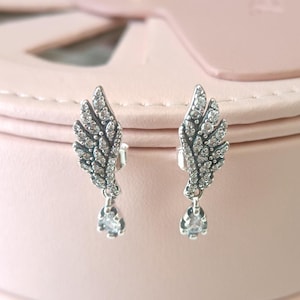 May include: A pair of silver dangle earrings with a wing design and clear crystal accents. The wings are detailed with smaller crystals and the earrings have a teardrop-shaped crystal dangling from the bottom.