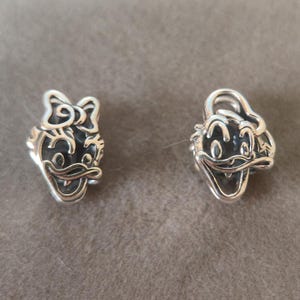 K&ouml;nnte beinhalten: Zwei silberne Charms mit Cartoon-Entengesichtern. Ein Charm hat eine Schleife, der andere einen Hut. Die Charms sind detailliert mit Gesichtsz&uuml;gen und sind wahrscheinlich f&uuml;r ein Armband oder eine Halskette.