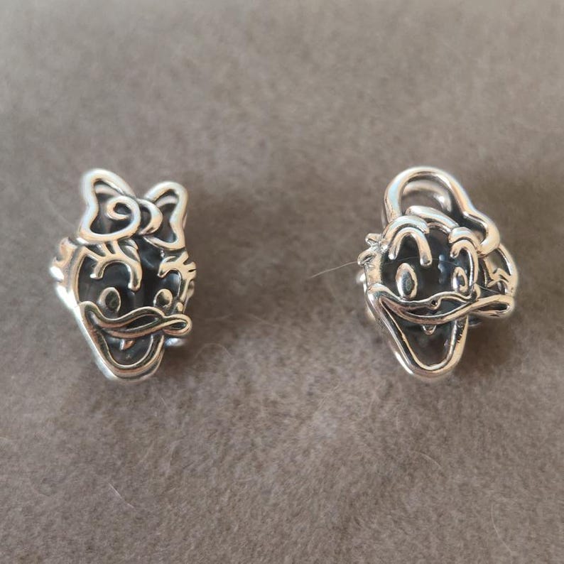 K&ouml;nnte beinhalten: Zwei silberne Charms mit Cartoon-Entengesichtern. Ein Charm hat eine Schleife, der andere einen Hut. Die Charms sind detailliert mit Gesichtsz&uuml;gen und sind wahrscheinlich f&uuml;r ein Armband oder eine Halskette.