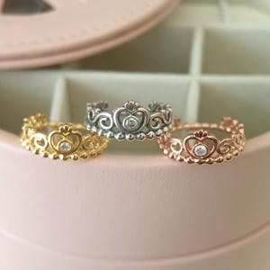 Può includere: Tre anelli a forma di corona da principessa in oro, argento e oro rosa, ognuno con un cristallo trasparente al centro della corona.