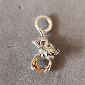 Puede incluir: Encanto de plata con un mono sosteniendo un plátano dorado. El mono está colgado boca abajo y tiene un bucle en la parte superior para sujetarlo a una pulsera o collar.