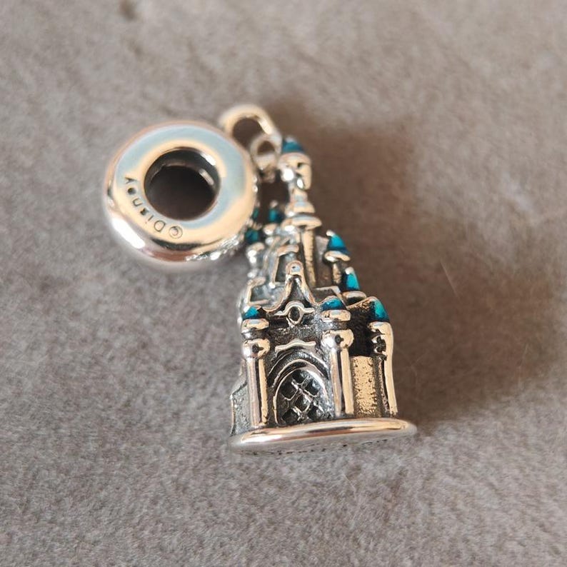 May include: Silver charm featuring a detailed miniature replica of a castle with blue enamel accents. The charm has a round bead with the word "Disney" engraved on it.