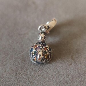 May include: A silver charm pendant with a round, multi-colored jewel-like design. The charm has a small loop at the top for attaching to a bracelet or necklace. The jewel-like design features various colors.