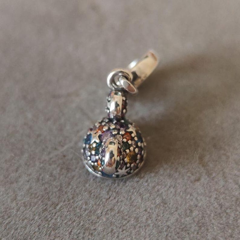 May include: A silver charm pendant with a round, multi-colored jewel-like design. The charm has a small loop at the top for attaching to a bracelet or necklace. The jewel-like design features various colors.