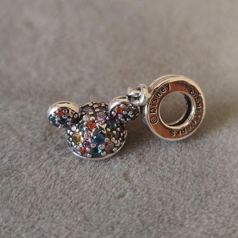 May include: A silver charm with a Mickey Mouse head design. The charm's head is encrusted with colorful gemstones. The charm has a ring with the word "Disney Parks" engraved on it.