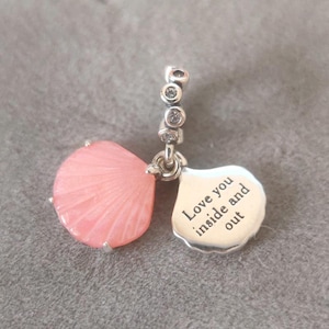 May include: A silver charm with a pink, petal-shaped pendant and a smaller, silver pendant with the words "Love you inside and out." The charm has a series of small, clear stones.