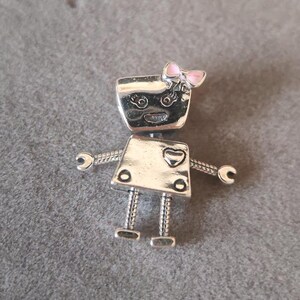 May include: Silver charm in the shape of a robot with a pink bow on its head. The robot has a heart on its chest and a cute face with eyes and a smile.