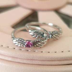 Op de afbeelding: Twee zilveren ringen met engelenvleugels. Een ring heeft een roze edelsteen en de andere een heldere edelsteen.