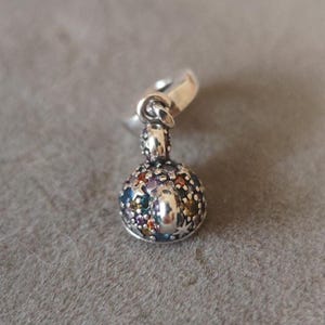 May include: A silver charm with a round, gem-studded design. The charm features a variety of colorful gemstones, including blue, green, purple, and yellow. The charm has a small loop at the top for attaching to a bracelet or necklace.