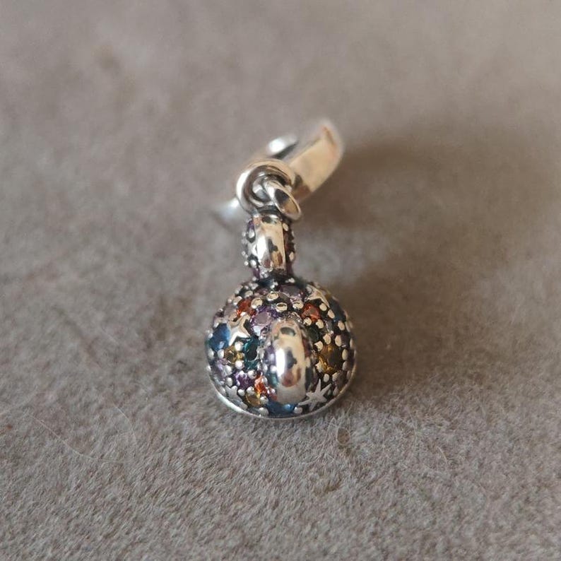 May include: A silver charm with a round, gem-studded design. The charm features a variety of colorful gemstones, including blue, green, purple, and yellow. The charm has a small loop at the top for attaching to a bracelet or necklace.
