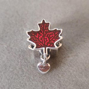 May include: A silver charm with a red maple leaf design and a small heart pendant. The heart pendant has the word "CANADA" engraved on it. The charm is likely made of sterling silver.