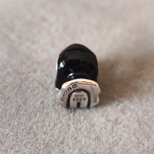 May include: A black and silver charm with the Disney logo and the text "S925". The charm has a rounded top and a silver base. The charm is likely made of sterling silver.