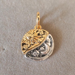 May include: A gold-tone pendant featuring a sun and moon design. The sun side is gold with a detailed sunburst and stars, while the moon side is silver with a crescent moon, stars, and a central gemstone. The pendant has a small loop for a chain.