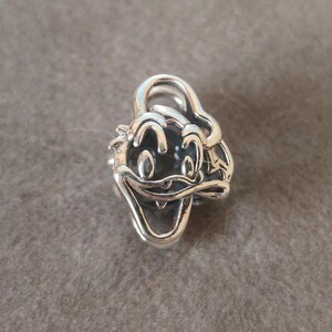 K&ouml;nnte beinhalten: Ein silberner Charm mit dem Gesicht von Donald Duck. Der Charm hat ein detailliertes Design mit schwarzem Hintergrund f&uuml;r Augen und Mund und ist wahrscheinlich aus Sterlingsilber gefertigt.