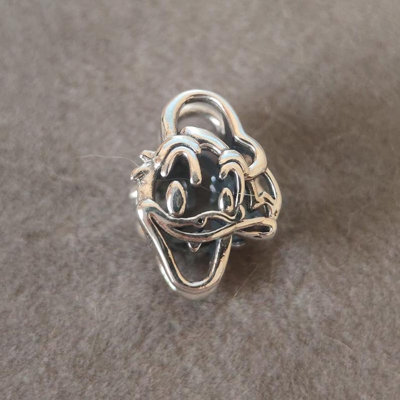 K&ouml;nnte beinhalten: Ein silberner Charm mit dem Gesicht von Donald Duck. Der Charm hat ein detailliertes Design mit schwarzem Hintergrund f&uuml;r Augen und Mund und ist wahrscheinlich aus Sterlingsilber gefertigt.