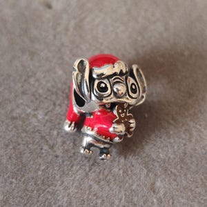 May include: A silver charm of a cartoon character in a red Santa hat and coat, holding a gingerbread man. The charm features large eyes and is set against a neutral background.
