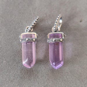 May include: A pair of amethyst crystal pendant earrings. Each earring has a light purple, elongated crystal with a silver decorative band and a silver hoop. The earrings are shown on a grey surface.