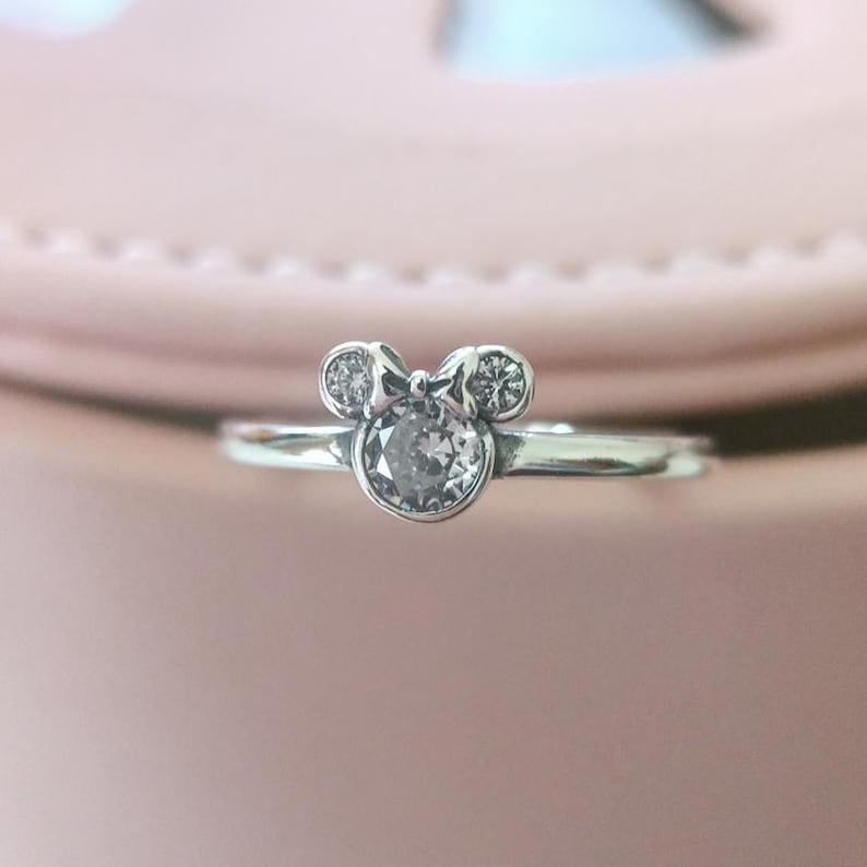 May include: A silver ring with a clear crystal in the center, shaped like Mickey Mouse's head with a bow.