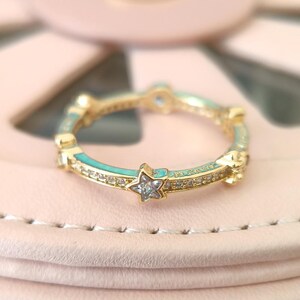 Puede incluir: Un anillo de oro con una banda de esmalte turquesa y peque&ntilde;os acentos de estrella engastados con piedras de circonita c&uacute;bica transparentes.