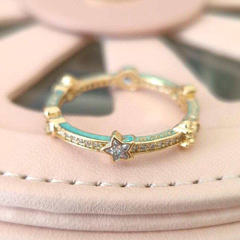 Puede incluir: Un anillo de oro con una banda de esmalte turquesa y peque&ntilde;os acentos de estrella engastados con piedras de circonita c&uacute;bica transparentes.