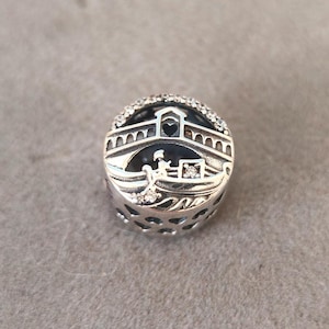 Może przedstawiać: Srebrny charm przedstawiający szczegółową scenę gondoli przepływającej pod mostem z wyciętym sercem. Charm jest ozdobiony małymi, przezroczystymi cyrkoniami.