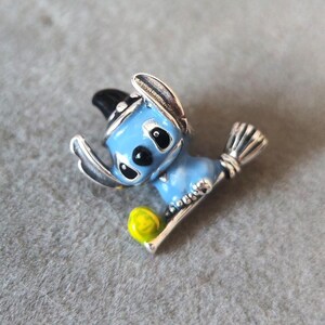 May include: A blue and silver-toned metal charm featuring a cartoon character with a black hat and a yellow ball. The charm is detailed with black eyes and a silver broom.