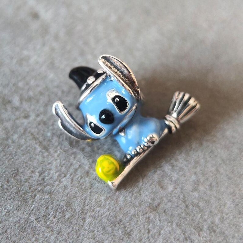 May include: A blue and silver-toned metal charm featuring a cartoon character with a black hat and a yellow ball. The charm is detailed with black eyes and a silver broom.