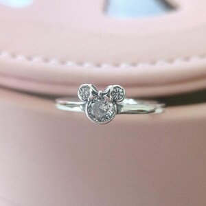 May include: A silver ring with a clear crystal in the center, shaped like Mickey Mouse's head with a bow.