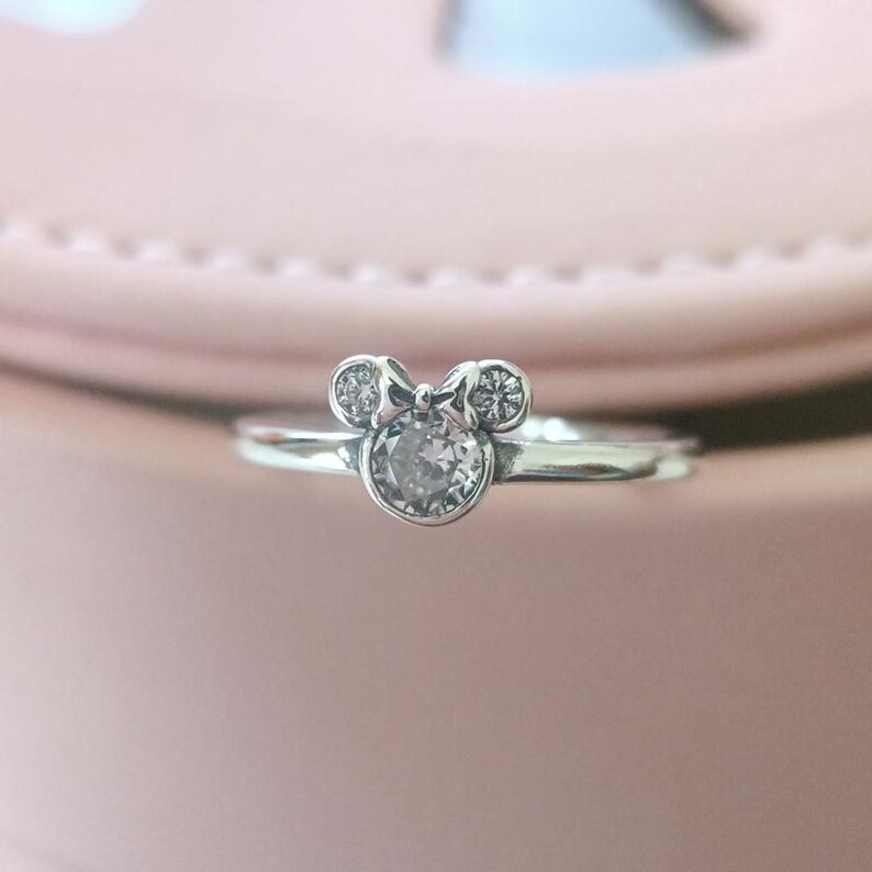 May include: A silver ring with a clear crystal in the center, shaped like Mickey Mouse's head with a bow.