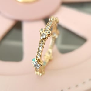 Puede incluir: Un anillo de oro con una fila de peque&ntilde;as piedras transparentes y un dise&ntilde;o de cabeza de Mickey Mouse con piedras transparentes. El anillo tambi&eacute;n tiene peque&ntilde;as piedras azules en forma de estrellas.