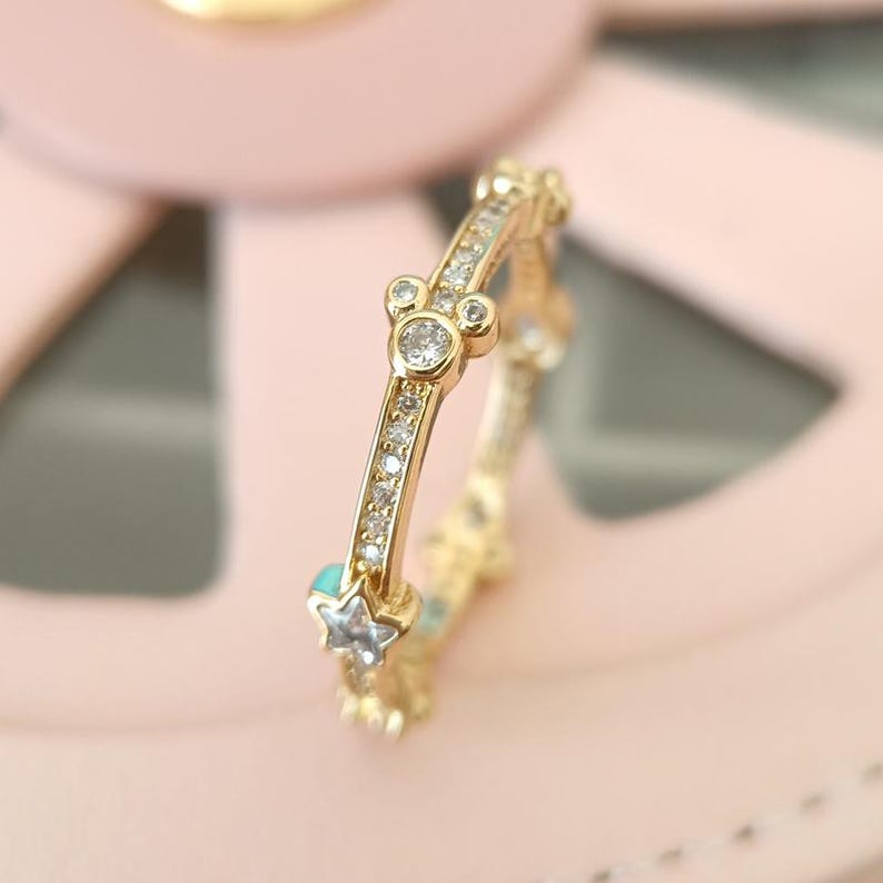 Puede incluir: Un anillo de oro con una fila de peque&ntilde;as piedras transparentes y un dise&ntilde;o de cabeza de Mickey Mouse con piedras transparentes. El anillo tambi&eacute;n tiene peque&ntilde;as piedras azules en forma de estrellas.