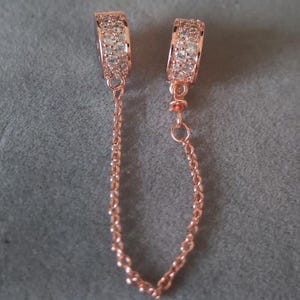 May include: Rose gold-toned jewelry safety chain with two decorative clasps. Each clasp is encrusted with small, clear crystals. A delicate chain connects the clasps, designed to secure jewelry.
