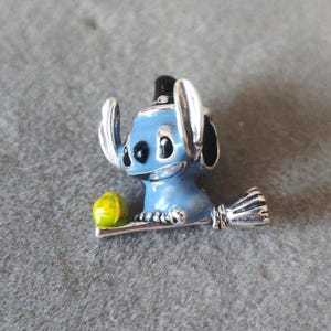 May include: A blue and silver charm featuring a cartoon character with large ears and a smiling face. The charm has a yellow and green object on the left side and a silver broom on the right. The charm is likely made of metal.