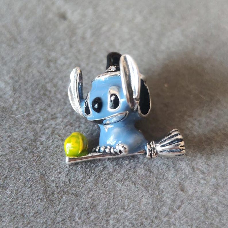 May include: A blue and silver charm featuring a cartoon character with large ears and a smiling face. The charm has a yellow and green object on the left side and a silver broom on the right. The charm is likely made of metal.