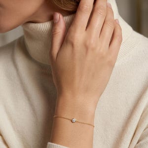 Op de afbeelding: Een delicate gouden kettingarmband met een enkele ronde, heldere, in een zetting geplaatste edelsteen in het midden. De armband is gemaakt van een fijne, gepolijste schakelketting die om de pols wordt vastgemaakt met een eenvoudige sluiting.