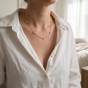 May include: A gold chain necklace featuring evenly spaced, round, clear stones set in metal bezels. The necklace rests against the collarbone, worn with a white button-down shirt. The design includes multiple small, circular, sparkling accents.