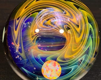 May include: A colorful glass marble with swirling patterns in shades of yellow, blue, and green. The marble features a central vortex design and a smaller, iridescent orb at the bottom. The overall effect is reminiscent of a galaxy.