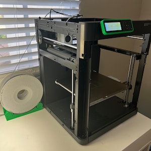 May include: A black 3D printer with a green display screen and a spool of filament on a white table.