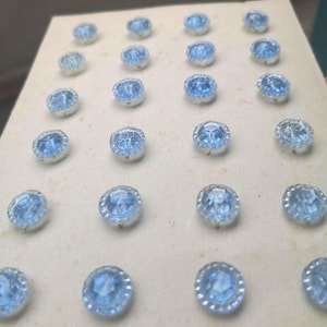 May include: A collection of light blue, faceted glass buttons with silver-colored metal rims. The buttons are arranged in neat rows on a cream-colored surface, showcasing their intricate design and reflective qualities. These buttons are ideal for sewing and craft projects.
