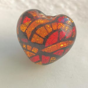 Puede incluir: Una cuenta de vidrio en forma de corazón con un diseño de vitral en tonos rojos, naranjas y amarillos. La cuenta tiene un acabado brillante y está decorada con purpurina.