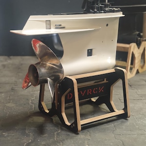 Könnte beinhalten: Ein Bootsmotor, überwiegend weiß, mit einem glänzenden silbernen Propeller und roten Akzenten. Er steht auf einem schwarz- und holzfarbenen Ständer mit dem roten Schriftzug "DVRCK". Der Motor ist vor einem dunklen Hintergrund platziert.