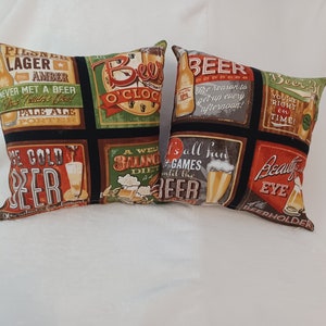 May include: Two decorative throw pillows with a black background and colorful beer-themed designs. The pillows feature various slogans and images related to beer, such as "Beer O'Clock", "It's all fun and games until the beer", and "Beauty is in the eye of the beerholder".