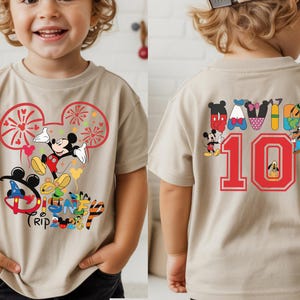 May include: A beige short-sleeved t-shirt with a Mickey Mouse design, fireworks, and the words "Disney Trip 2023". The back features the number 10 in red. The shirt has a colourful design.