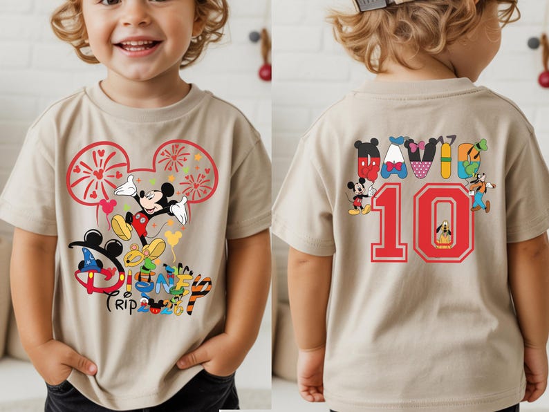 May include: A beige short-sleeved t-shirt with a Mickey Mouse design, fireworks, and the words "Disney Trip 2023". The back features the number 10 in red. The shirt has a colourful design.