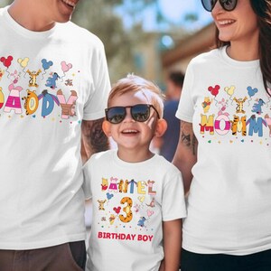 K&ouml;nnte beinhalten: Wei&szlig;e T-Shirts mit farbenfrohen Designs zum Thema Winnie Puuh. Ein Shirt tr&auml;gt die Aufschrift "DADDY", ein anderes "MOMMY" und das dritte "DANIEL 3 BIRTHDAY BOY". Die Shirts zeigen Zeichentrickfiguren und Luftballons.