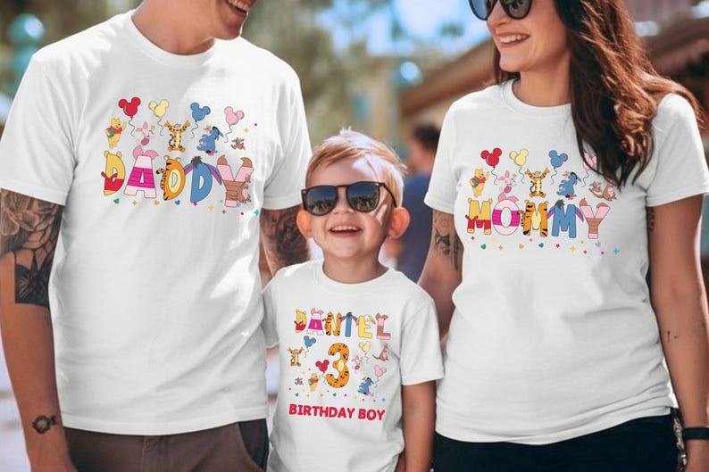 K&ouml;nnte beinhalten: Wei&szlig;e T-Shirts mit farbenfrohen Designs zum Thema Winnie Puuh. Ein Shirt tr&auml;gt die Aufschrift "DADDY", ein anderes "MOMMY" und das dritte "DANIEL 3 BIRTHDAY BOY". Die Shirts zeigen Zeichentrickfiguren und Luftballons.