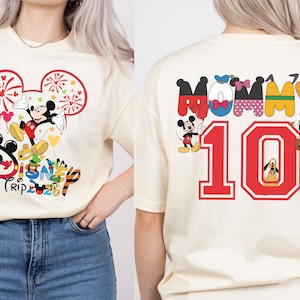 May include: Cream-coloured t-shirt with a Mickey Mouse graphic and the text "Disney Trip 2023" on the front. The back of the shirt shows the word "Mommy" and the number "10" with Disney character graphics. A fun, colourful design.